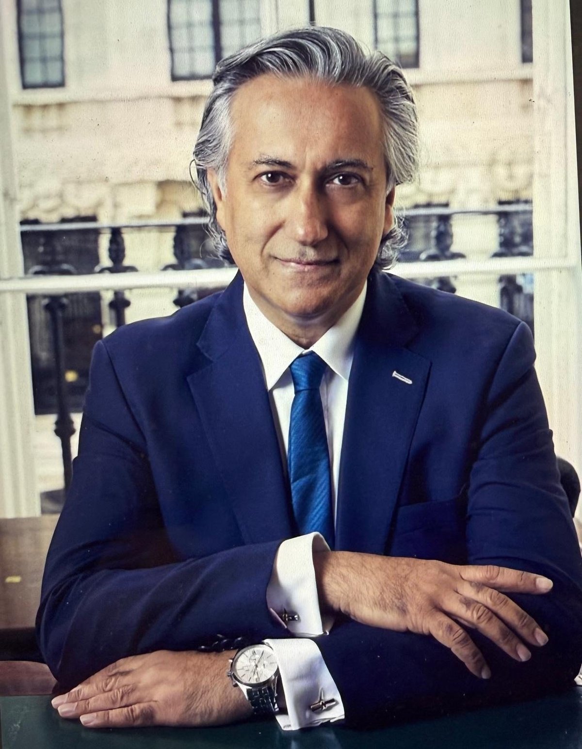 Professional headshot of a middle-aged man in dark blue suit and tie with gray hair, sitting at desk with classical building backdrop